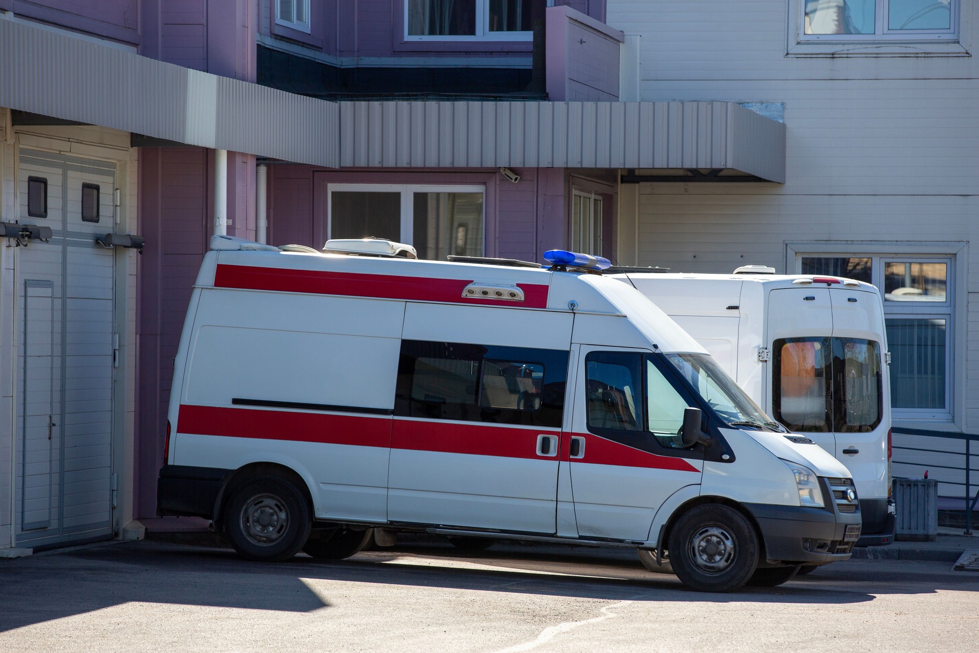 The image shows an ambulance parked outside a building. The vehicle is white with a red stripe and has emergency lights on top.